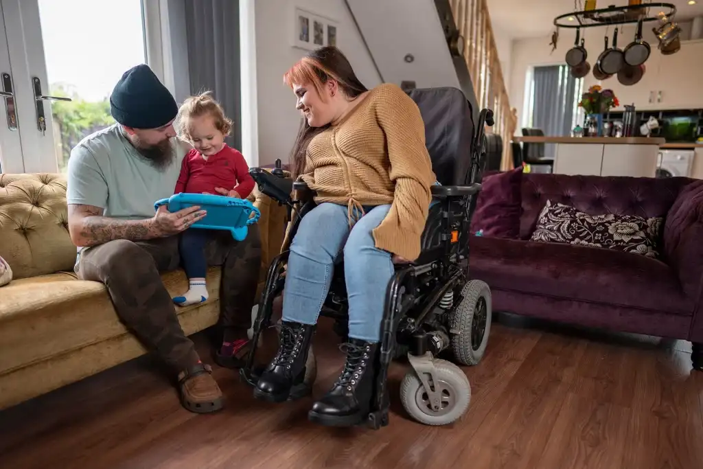 Mother in a power wheelchair spending quality time with her partner and child in a warm, accessible home environment.
