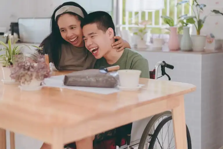 Happy young person in a wheelchair sharing a joyful moment with a caregiver at home, representing NDIS support and compassionate care.